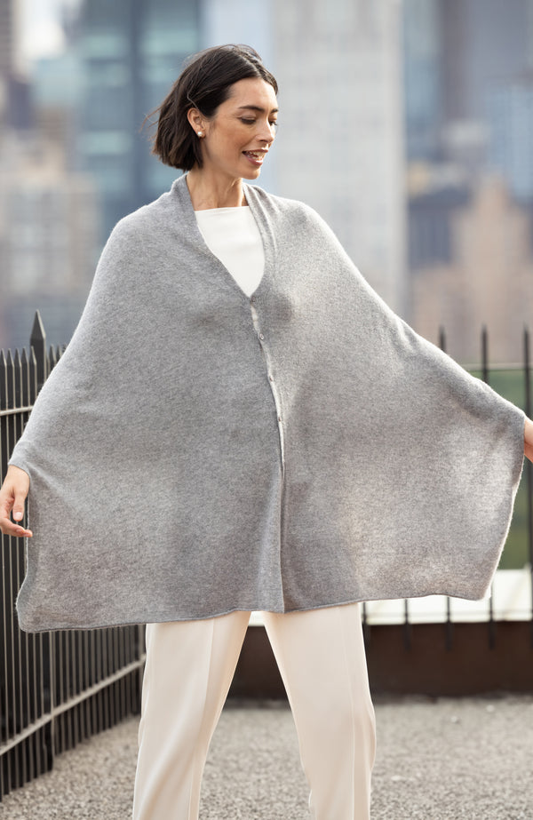 Woman wearing a CASHMERE BUTTON SHOULDER PONCHO + WRAP in medium grey with a blurred cityscape background